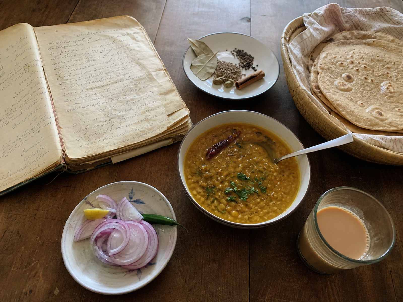 A recipe notebook beside dal, roti, spices, and chai on a wooden table